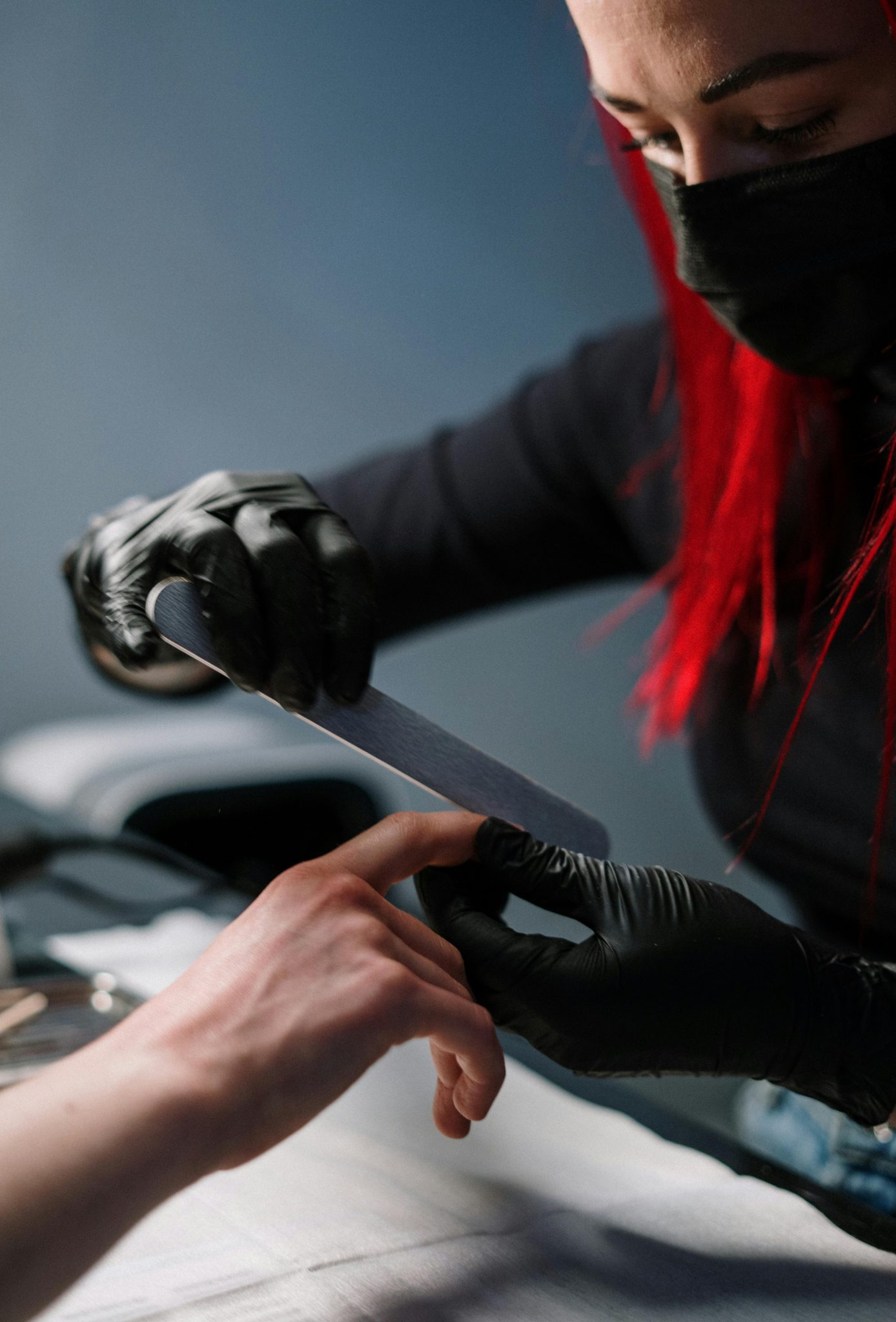 A red-haired technician filing nails at a salon, wearing black gloves and face mask.