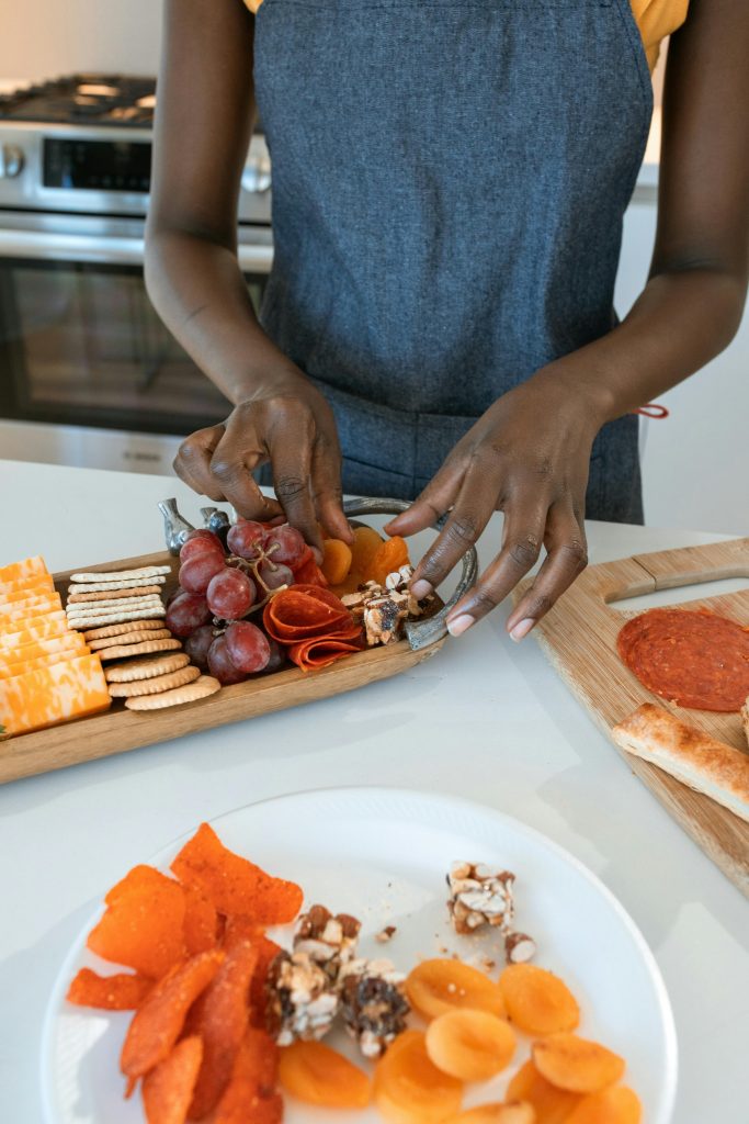 Person arranging a variety of cheeses, meats, and fruits on a charcuterie board indoors.